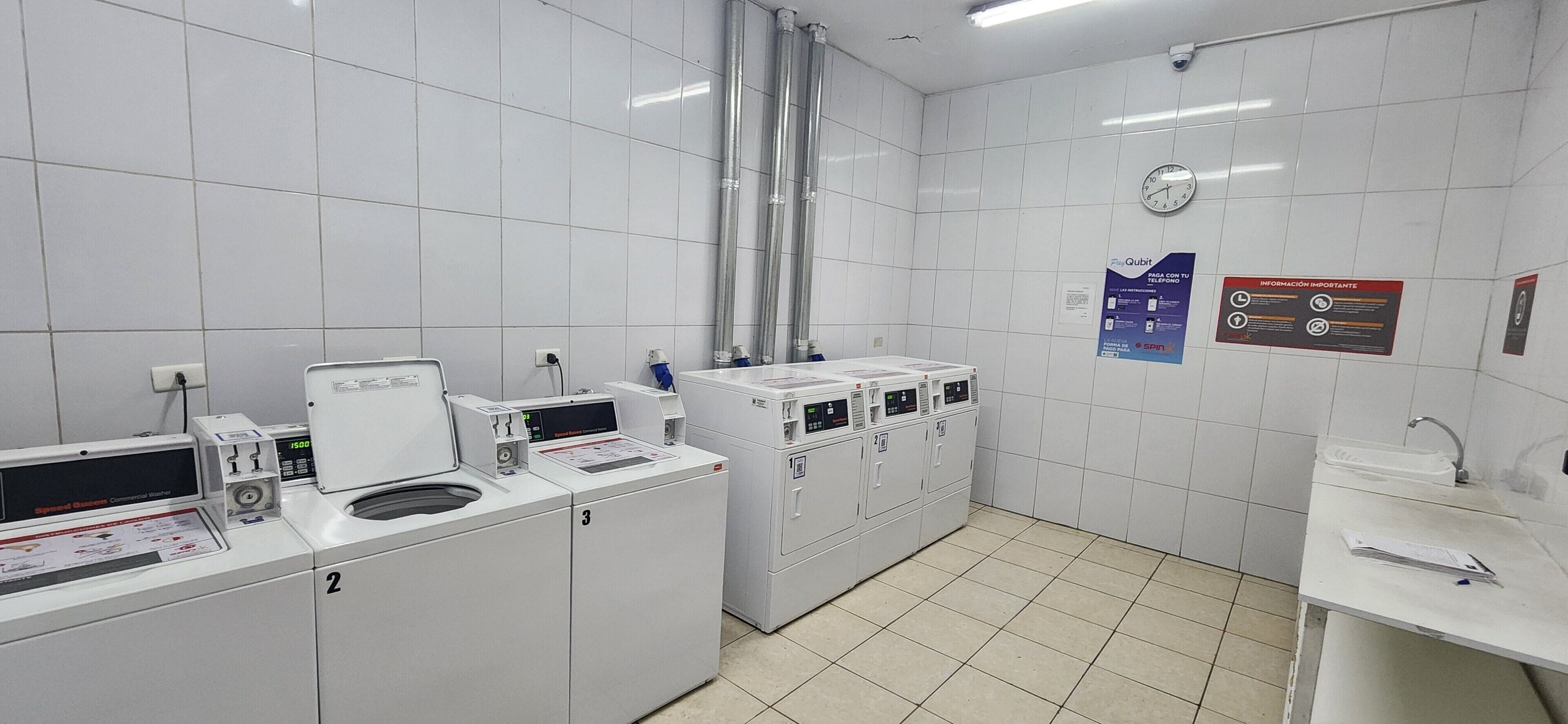 A clean, well-lit laundry facility with multiple washing machines and dryers, a clock, and informational posters on the walls.