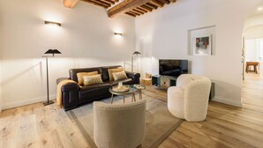 Un soggiorno moderno con un divano in pelle nera, una poltrona beige e un tavolino da caffè in vetro. Presente una TV a schermo piatto montata a parete, e la stanza è caratterizzata da travi a vista sul soffitto.