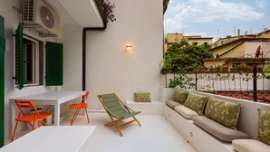Un balcone con un tavolo bianco, due sedie arancioni, una poltrona da relax verde e un divano con cuscini verdi.
