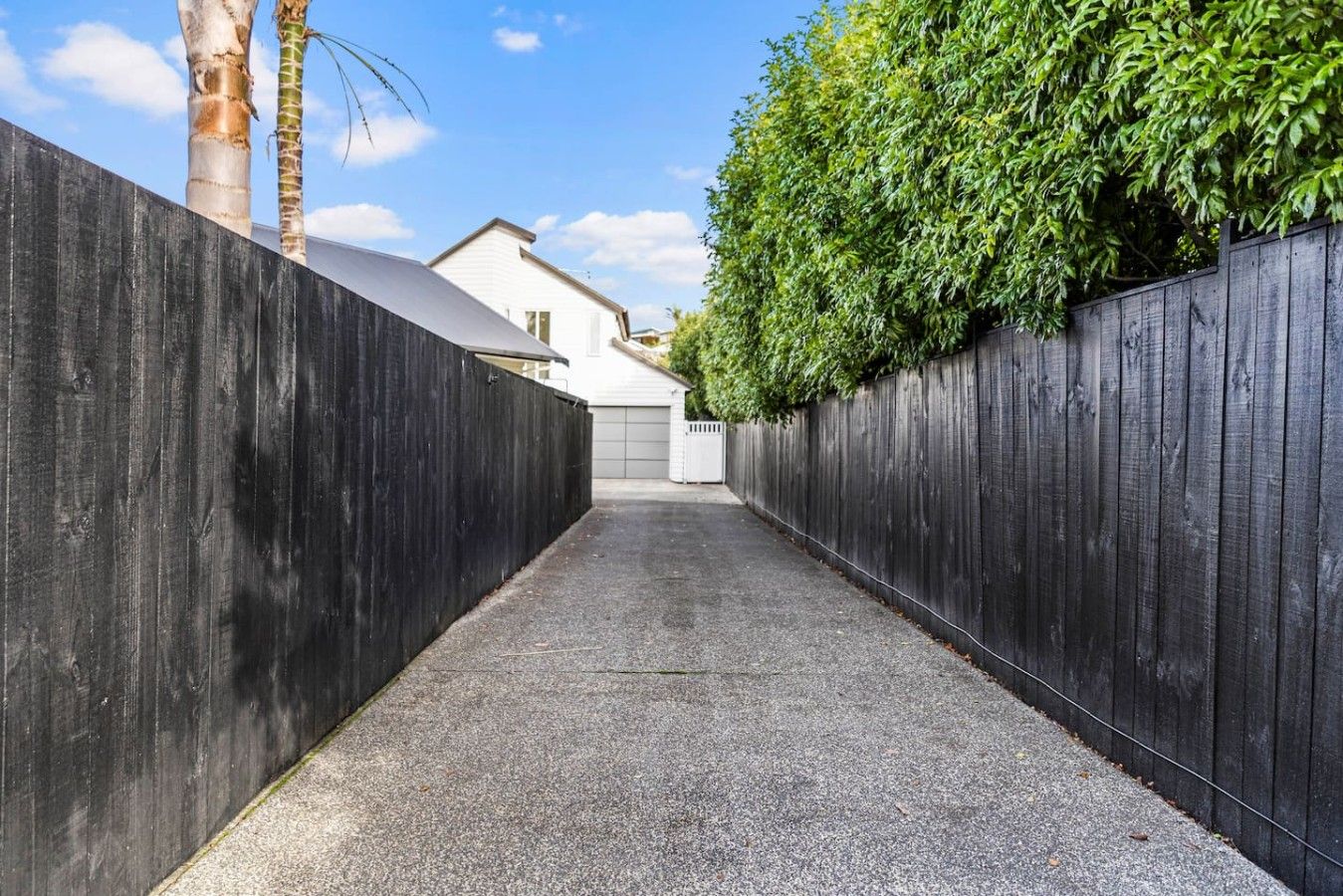 A narrow paved pathway flanked by dark wooden fences, leading to a white building with a garage.