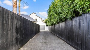 Un étroit sentier pavé, bordé de clôtures en bois sombre, mène à un bâtiment blanc doté d’un garage.