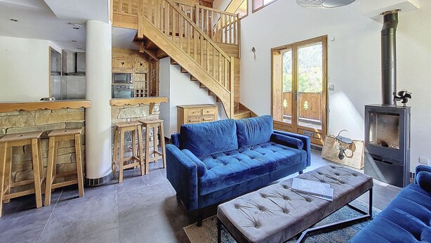 A spacious living area with a blue sofa, a wooden staircase, a kitchen area, and a wood-burning stove. - APARTMENT BALCONS DU MONT D'EVIAN - 8 PEOPLE - ST JEAN D'AULPS VILLAGE (Saint-Jean-d´Aulps)