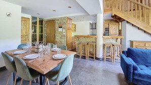 A dining area with a wooden table set for four, surrounded by chairs. A kitchen with wooden cabinets and a stone wall is visible in the background. - APARTMENT BALCONS DU MONT D'EVIAN - 8 PEOPLE - ST JEAN D'AULPS VILLAGE (Saint-Jean-d´Aulps)