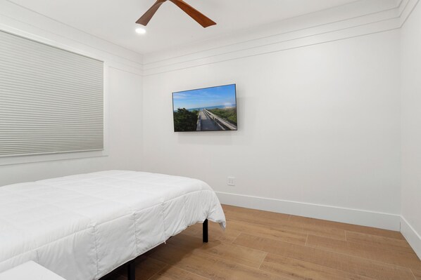 Quarto com cama branca, piso de madeira, janela com persianas e quadro com uma estrada na parede.