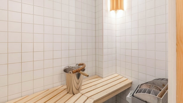 Un baño con paredes de azulejos blancos, un banco de madera, un cubo de metal y un perchero para toallas.