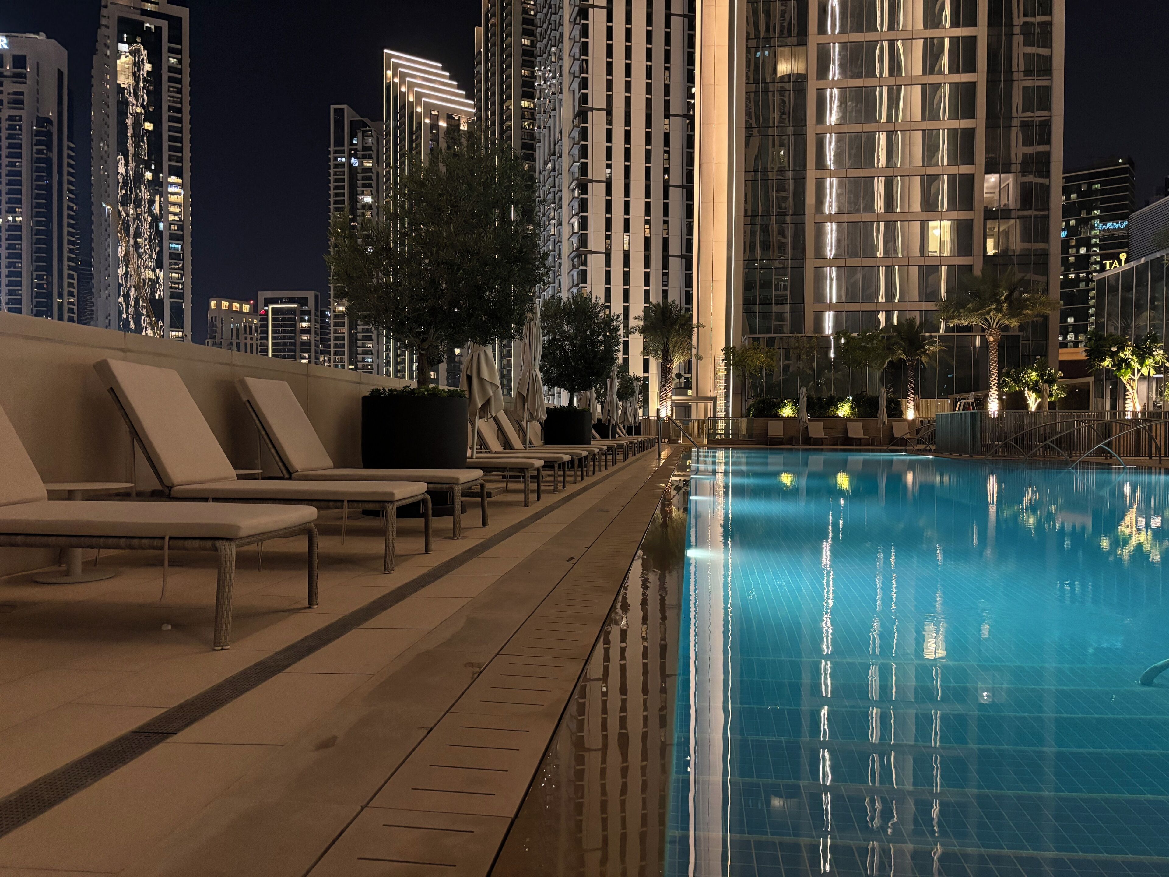 A nighttime pool area with lounge chairs and a swimming pool, surrounded by high-rise buildings.