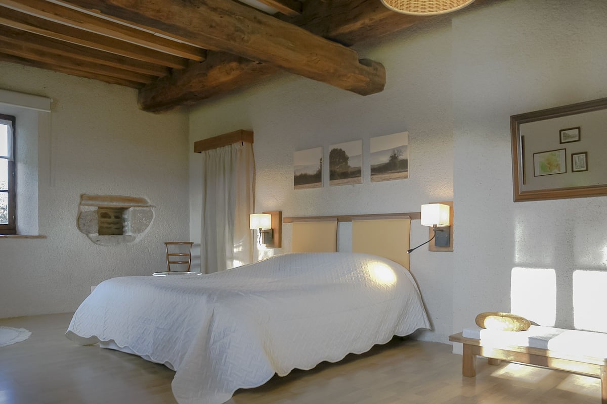 Un dormitorio con una cama grande, vigas de madera en el techo, una mesita de noche con lámpara y cuadros enmarcados en la pared.
