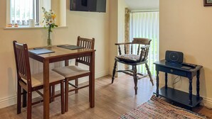Un comedor con una mesa y sillas de madera, un televisor montado en la pared, y una mesita con un altavoz y control remoto.