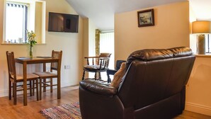 Una sala de estar con un sofá de cuero marrón, una mesa de comedor de madera con sillas, un televisor montado en la pared y una ventana con cortinas.