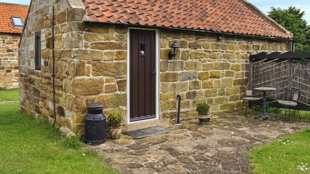 Una cabaña de piedra con techo de tejas, puerta de madera y un pequeño jardín con macetas y muebles de exterior.
