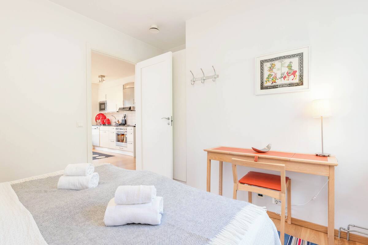 A hotel room with a bed, a desk, and a kitchen visible through an open door.