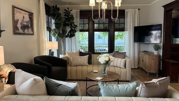 A living room with a sofa, armchairs, a coffee table, and a TV. There is a large window with curtains, a plant, and a painting on the wall. - Historic Elegance in Prime Location! (Milwaukee)