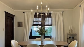 A dining area with a wooden table, six chairs, a chandelier, a window with curtains, and a small shelf with candles. - Historic Elegance in Prime Location! (Milwaukee)