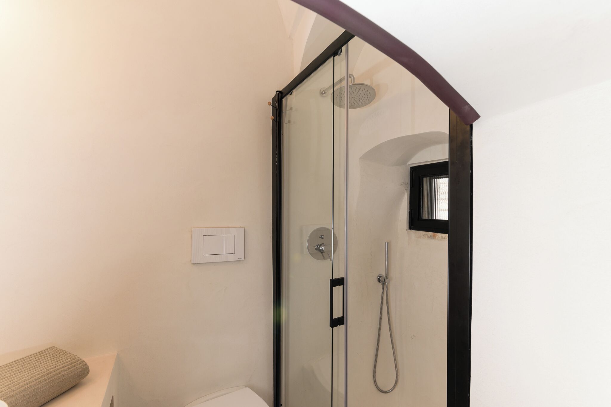 A glass-enclosed shower with a handheld showerhead, a wall-mounted light switch, and a small window.