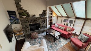 Ein gemütliches Wohnzimmer mit Kamin aus Stein, einem roten Sofa, einem weißen Sessel und einem großen Fenster mit Jalousien.