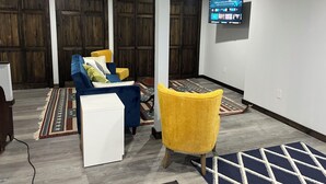 Ein Wohnzimmer mit einem blauen Sofa, einem gelben Sessel und einem weißen Schrank. Ein Fernseher ist an der Wand befestigt und im Hintergrund ist eine Holztür zu sehen.