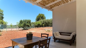 Une terrasse extérieure avec une table et des chaises en bois, un banc rembourré et une vue sur un court de tennis et des arbres.