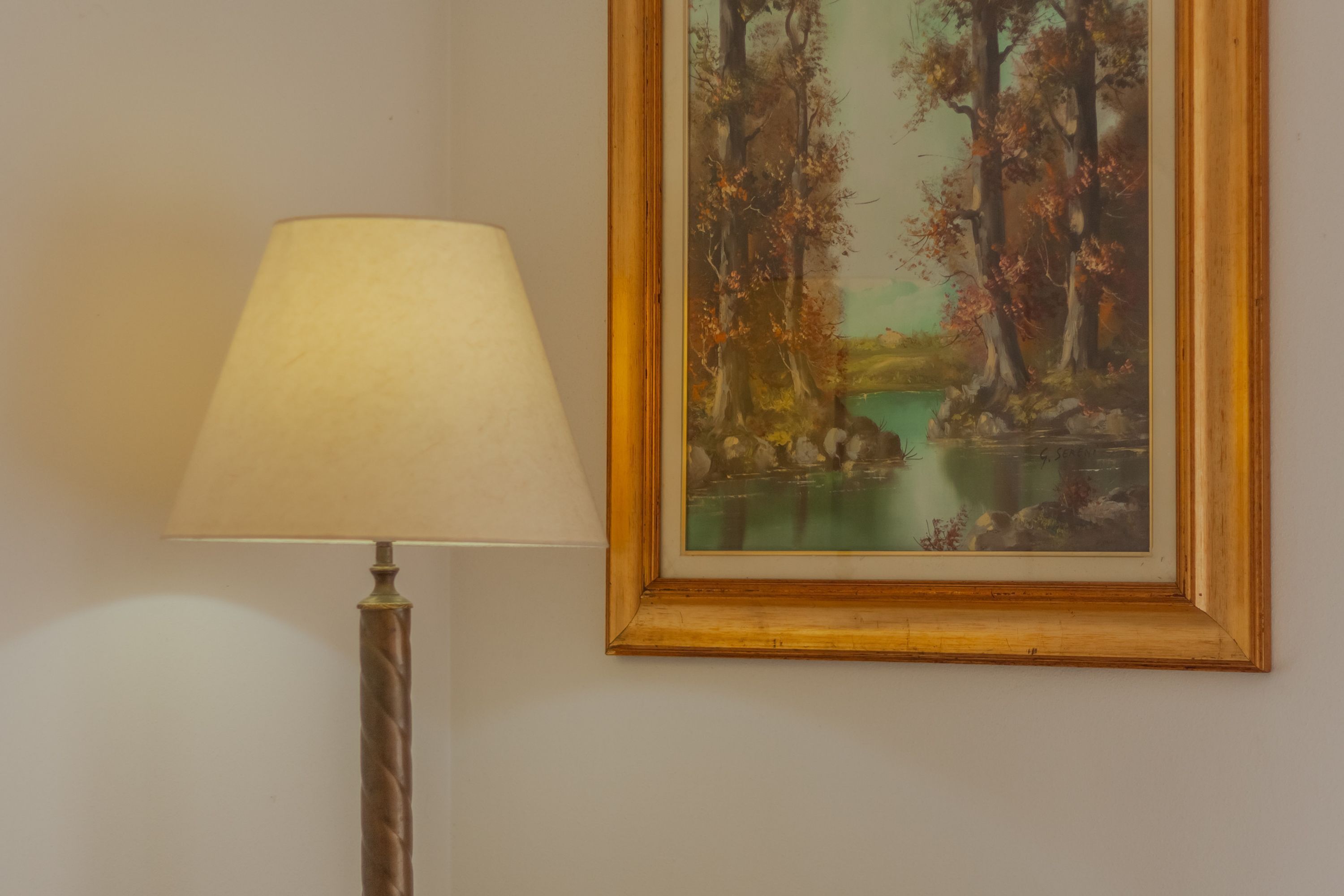 A lamp with a beige lampshade and a painting of a forest scene with a body of water.