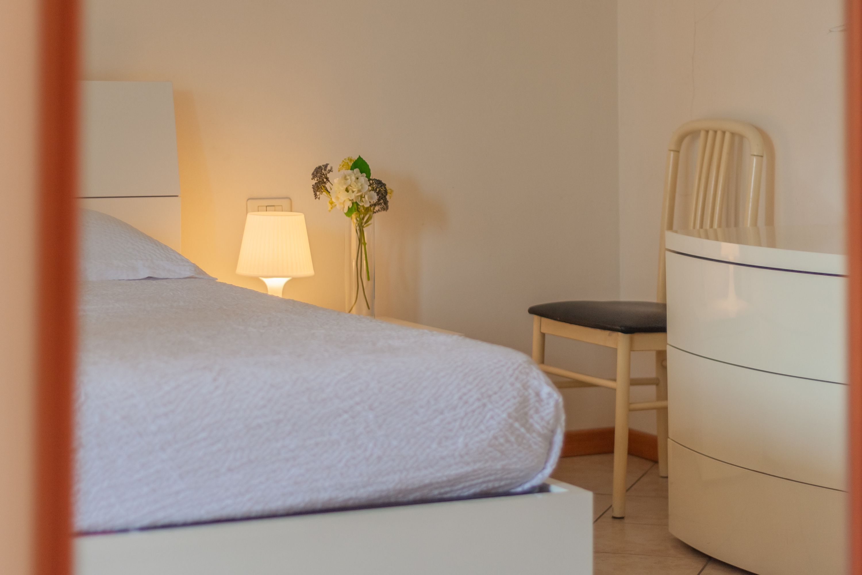 Un lit bien fait, recouvert d’une couverture blanche, une lampe de chevet, une chaise et une commode.