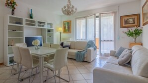 Una sala de estar moderna con comedor, muebles blancos, una araña y un amplio ventanal con cortinas.