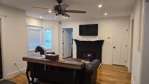 Ein Wohnzimmer mit Kamin, einem flachbildfernseher darüber, einem Deckenventilator und einem Essbereich mit Tisch und Stühlen.