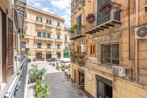 Una calle estrecha flanqueada por edificios, con balcones adornados con plantas en macetas, un café y un letrero de bar.