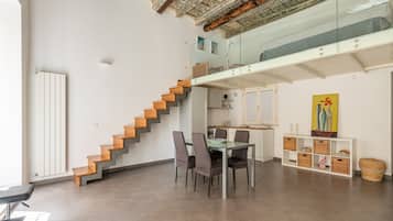 Um interior moderno com uma escada que leva a um loft, uma área de jantar com mesa de vidro e uma cozinha com armários brancos.
