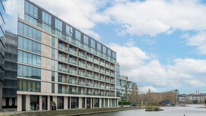 A modern multi-story building with a glass facade, a waterfront view, and a series of steps leading to the entrance. - Deluxe 1 Bedroom Apartment in City Road (London)