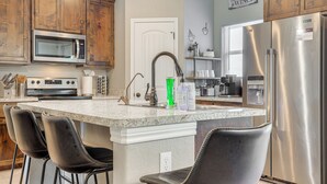 A kitchen with wooden cabinets, a stainless steel refrigerator, a microwave, a sink, and a bar area with two stools.