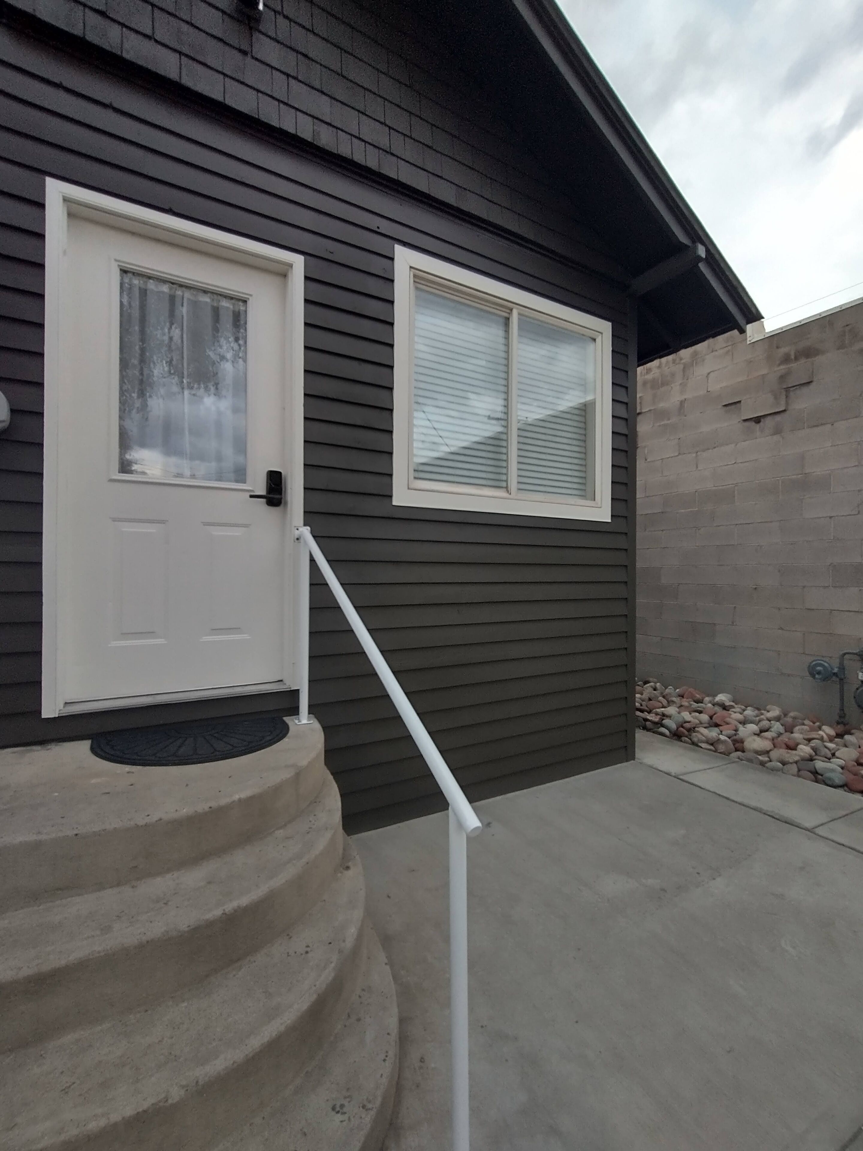 A residential building with a dark exterior, a white front door, a small window, and a concrete staircase leading to the entrance.