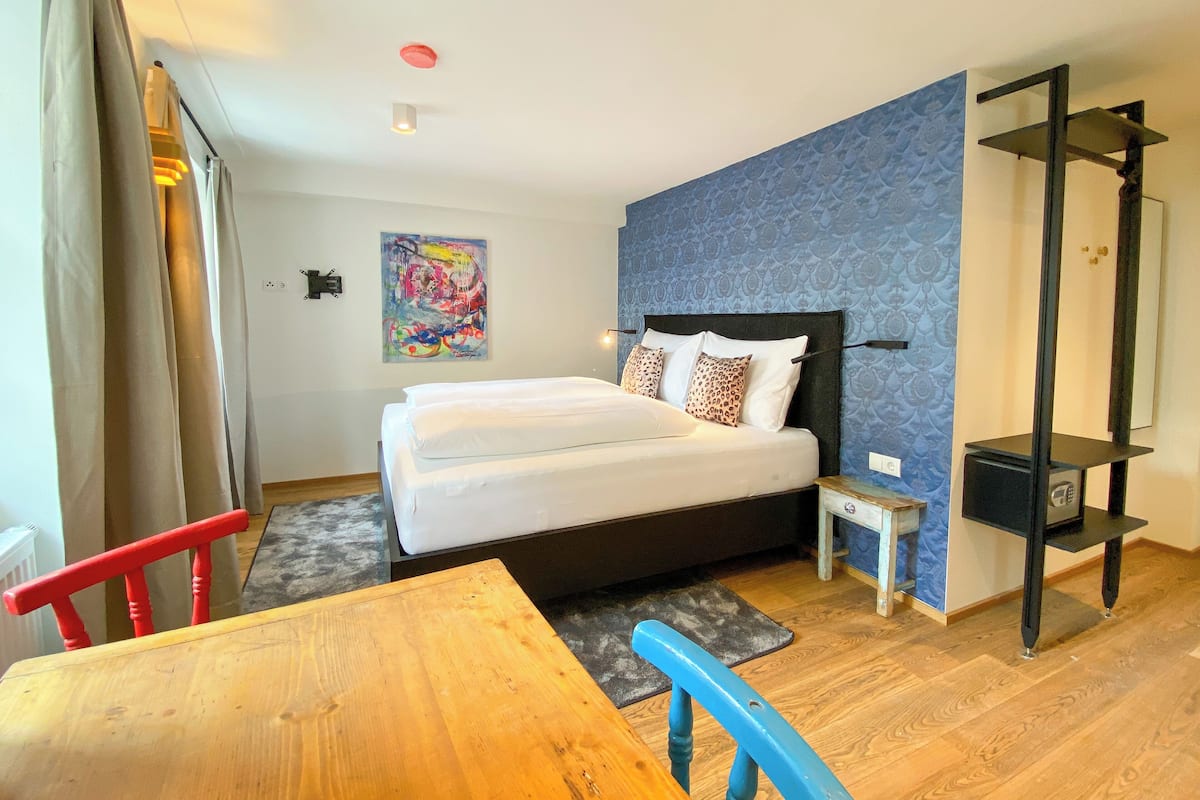 Una habitación de hotel con una cama, una mesita de noche pequeña, una silla, una mesa de madera, un muro con estampado azul y una lámpara de techo roja.