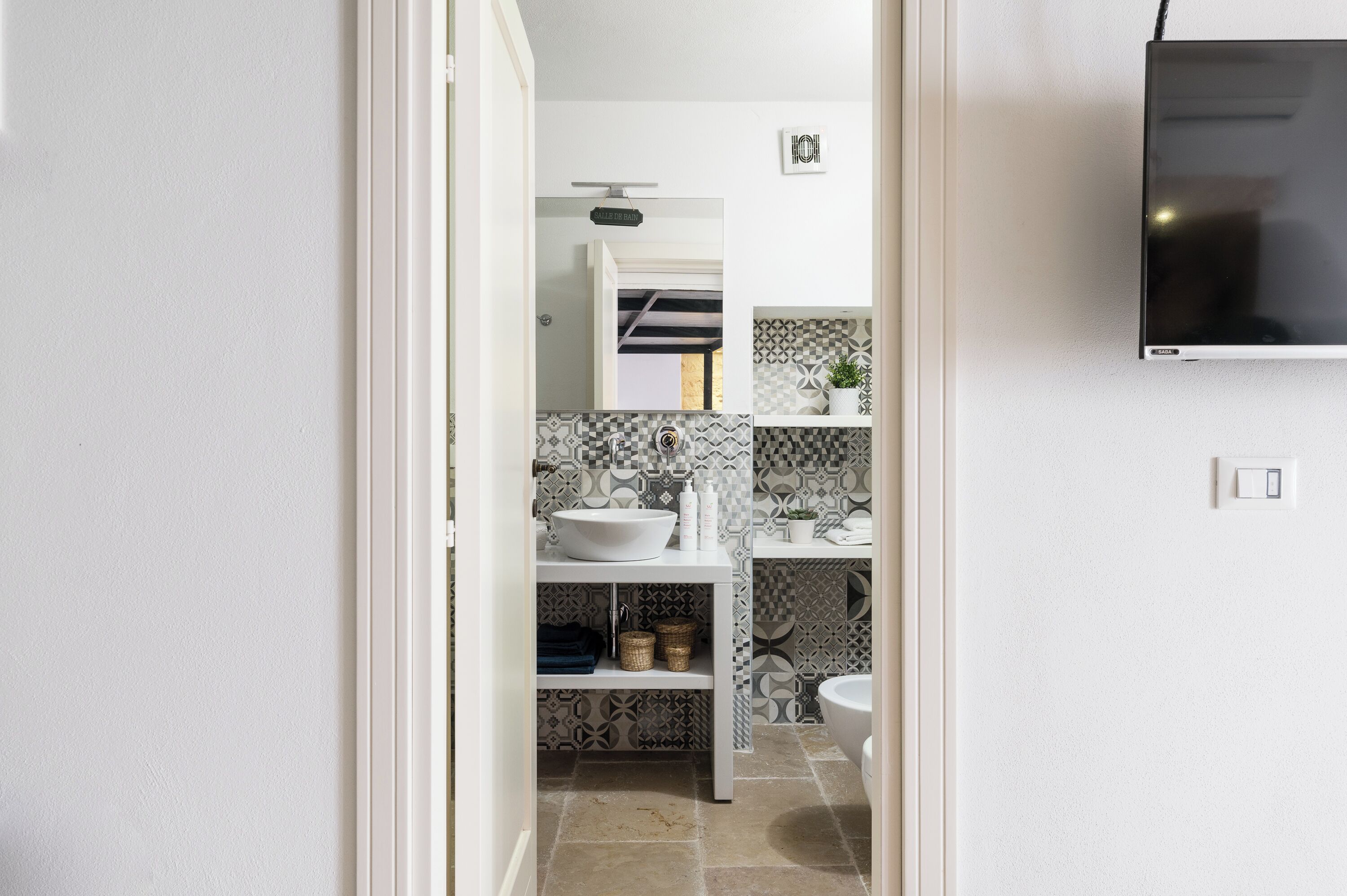 Une salle de bain moderne avec un lavabo blanc, un miroir et des toilettes. On y trouve une étagère avec des articles de toilette et une plante en pot.