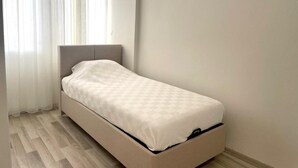 A small, single-bed room with a wooden floor, a plain white wall, and a window with sheer curtains.