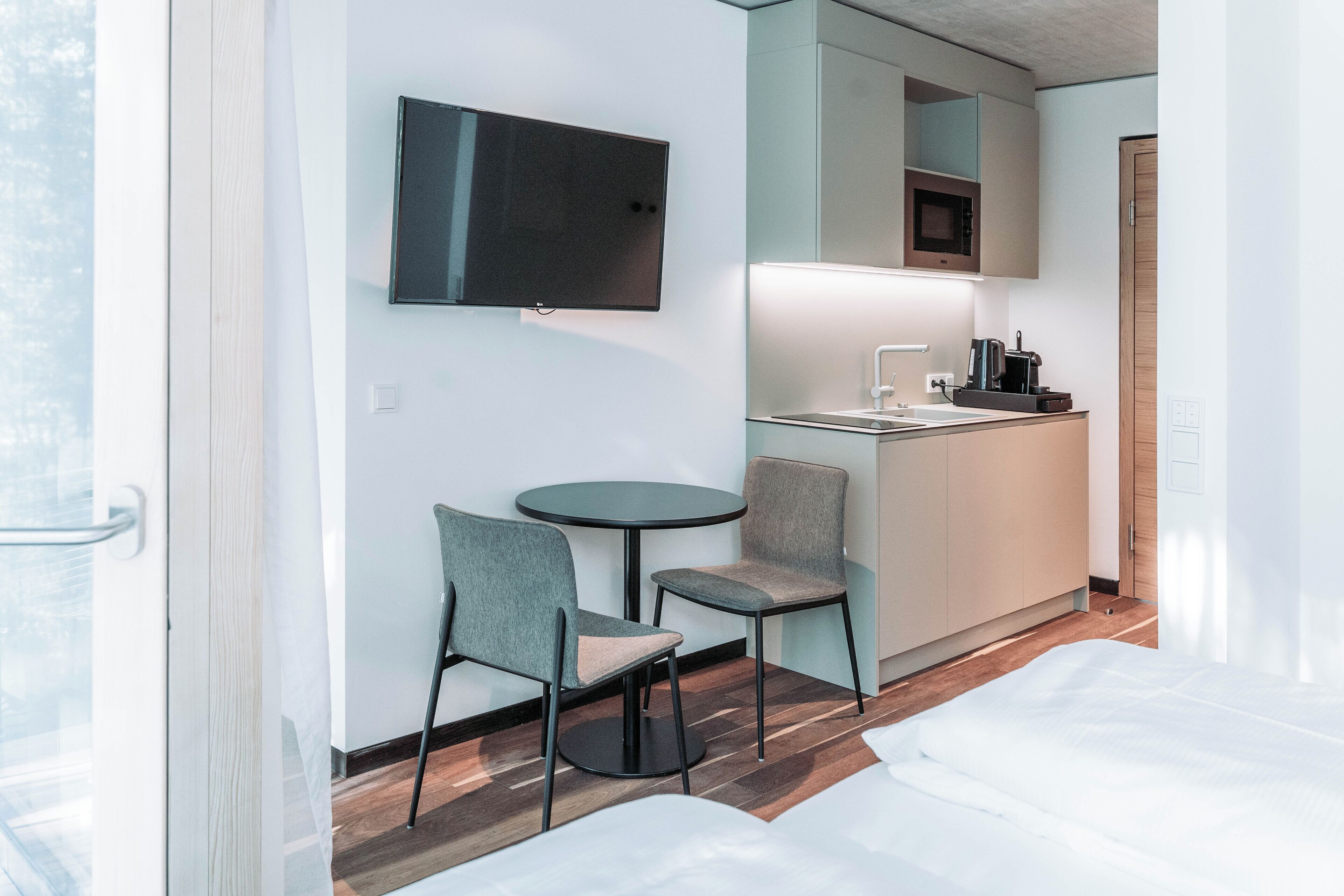 Quarto de hotel com cama, uma pequena mesa redonda com duas cadeiras, cozinha compacta e televisão na parede.