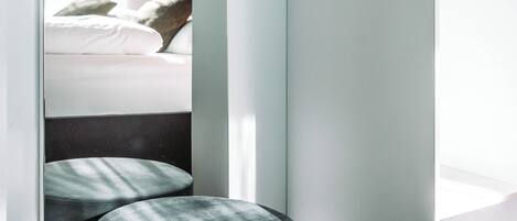 A modern hotel room with a bed, a small round ottoman, and a view of another room.