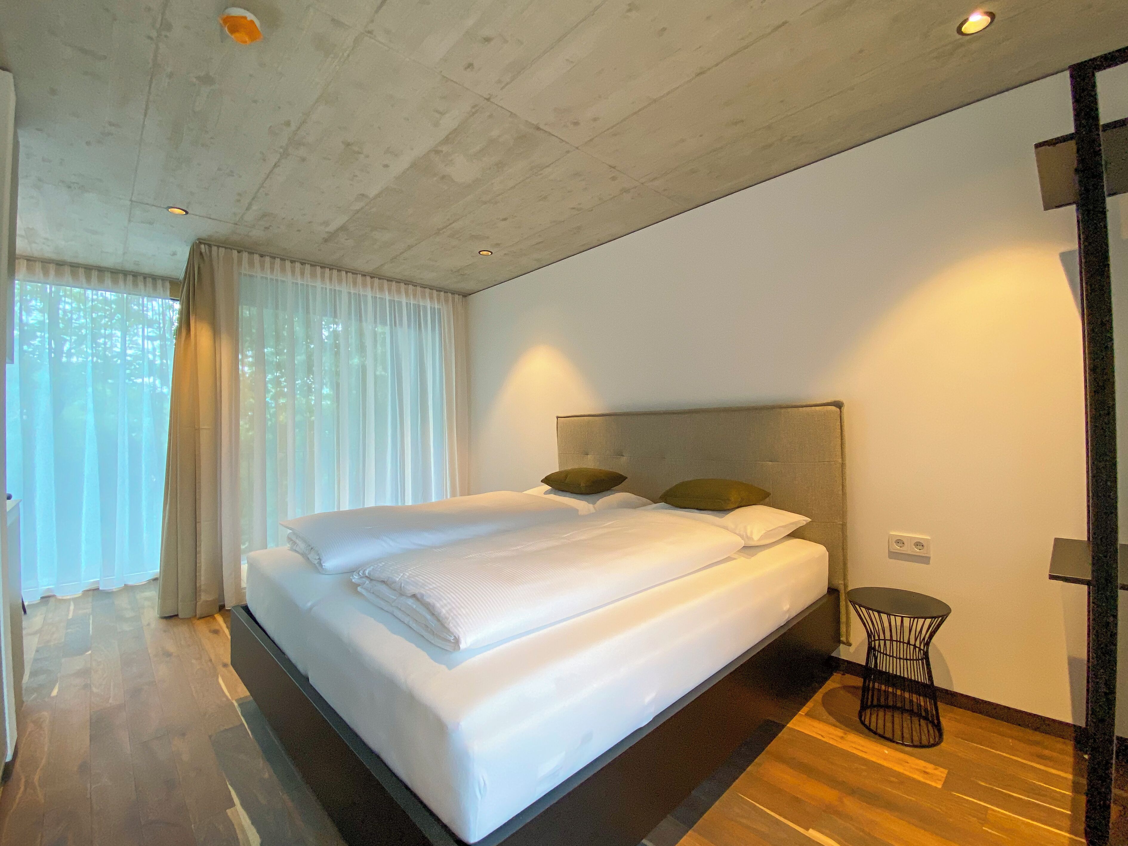 Une chambre d’hôtel moderne, dotée d’un grand lit, d’une petite table ronde et offrant une vue sur l’extérieur à travers de fines rideaux.