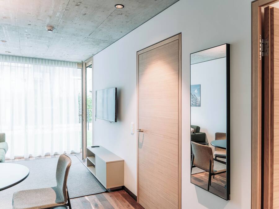 A modern hotel room with a large window, a flat-screen TV, a wooden door, and a dining area with a table and chairs.