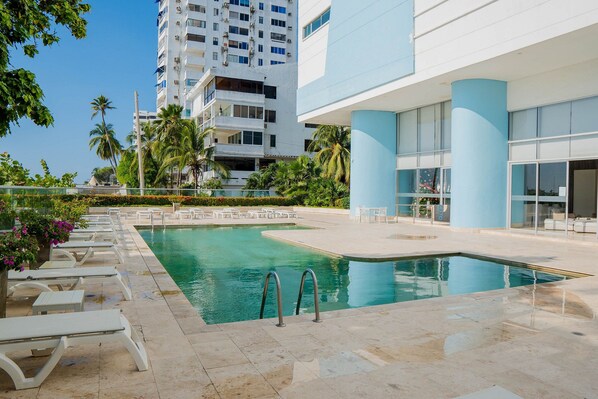 A swimming pool with lounge chairs and a modern building with large windows and columns. - U-GO Palmetto Sunset Junto al Mar (Cartagena)
