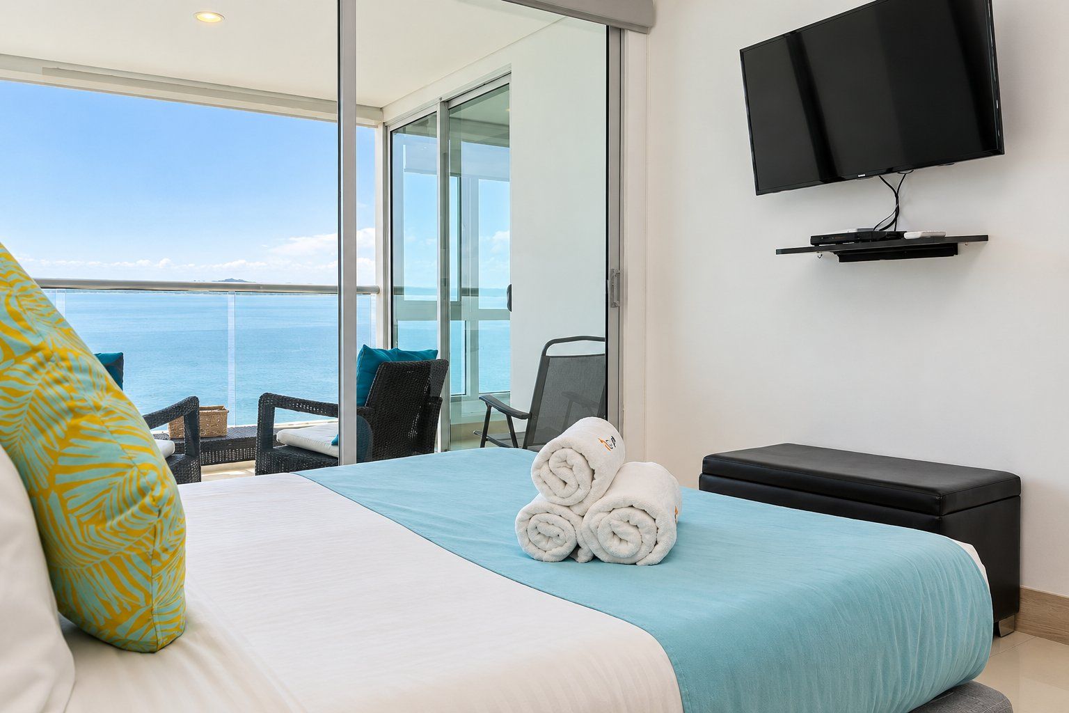 A hotel room with a bed, a TV, a view of the sea, and a balcony with chairs.