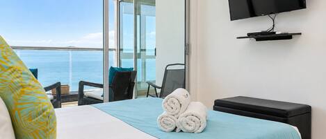 A hotel room with a bed, a TV, a view of the sea, and a balcony with chairs.