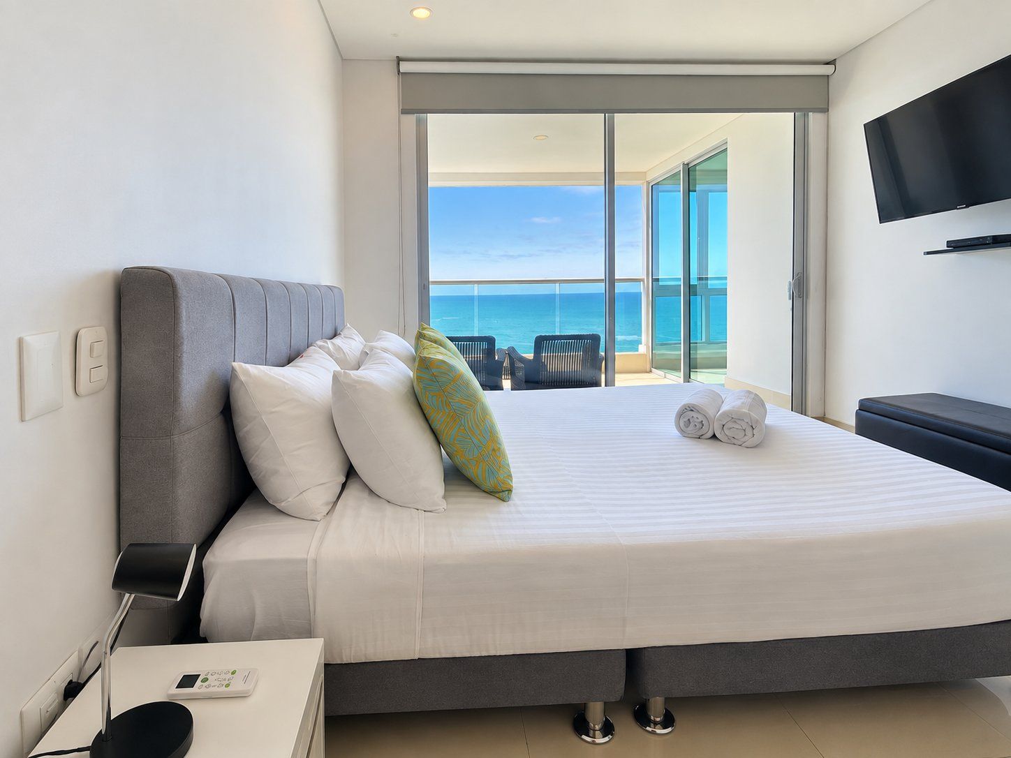 A hotel room with a bed, a TV, a desk with a lamp, and a view of the ocean.
