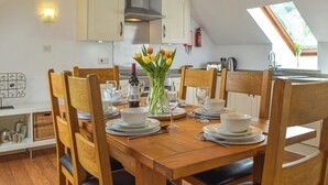 Un comedor con una mesa de madera dispuesta para cuatro personas, rodeada de sillas de madera. Un jarrón con tulipanes está sobre la mesa, y se ven electrodomésticos de cocina al fondo.