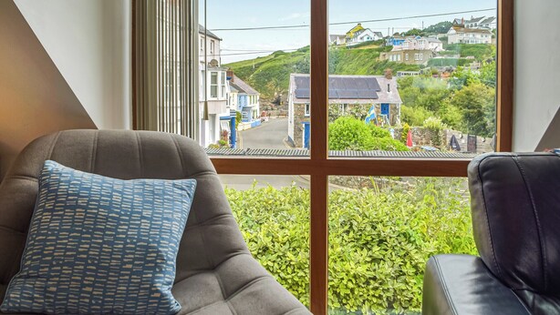 Una habitación con un amplio ventanal que ofrece vistas a una zona residencial, un sofá gris con un cojín estampado y un sillón de cuero negro.