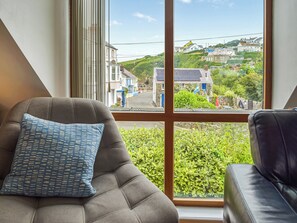 Ein Zimmer mit großem Fenster und Blick auf eine Wohngegend, ein graues Sofa mit gemustertem Kissen sowie ein schwarzer Ledersessel.