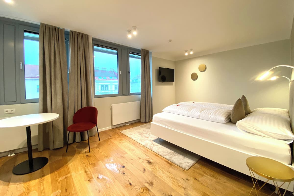 Una habitación de hotel moderna con una cama, una mesita pequeña, una silla roja y ventanas amplias.