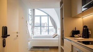 A compact hotel room with a bed, a kitchenette, and a large window offering a view of the cityscape. - Blackhome Wien (Vienna)