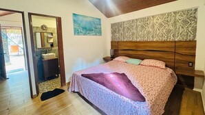 A bedroom with a wooden ceiling, a bed with patterned bedding, a nightstand with a lamp, a bathroom with a sink and mirror, and a door leading outside.