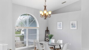 A dining area with a round table, white chairs, a chandelier, and a large window with blinds. - Hello Summer, full refund*, pool heat, sleeps 6 (Rotonda West)