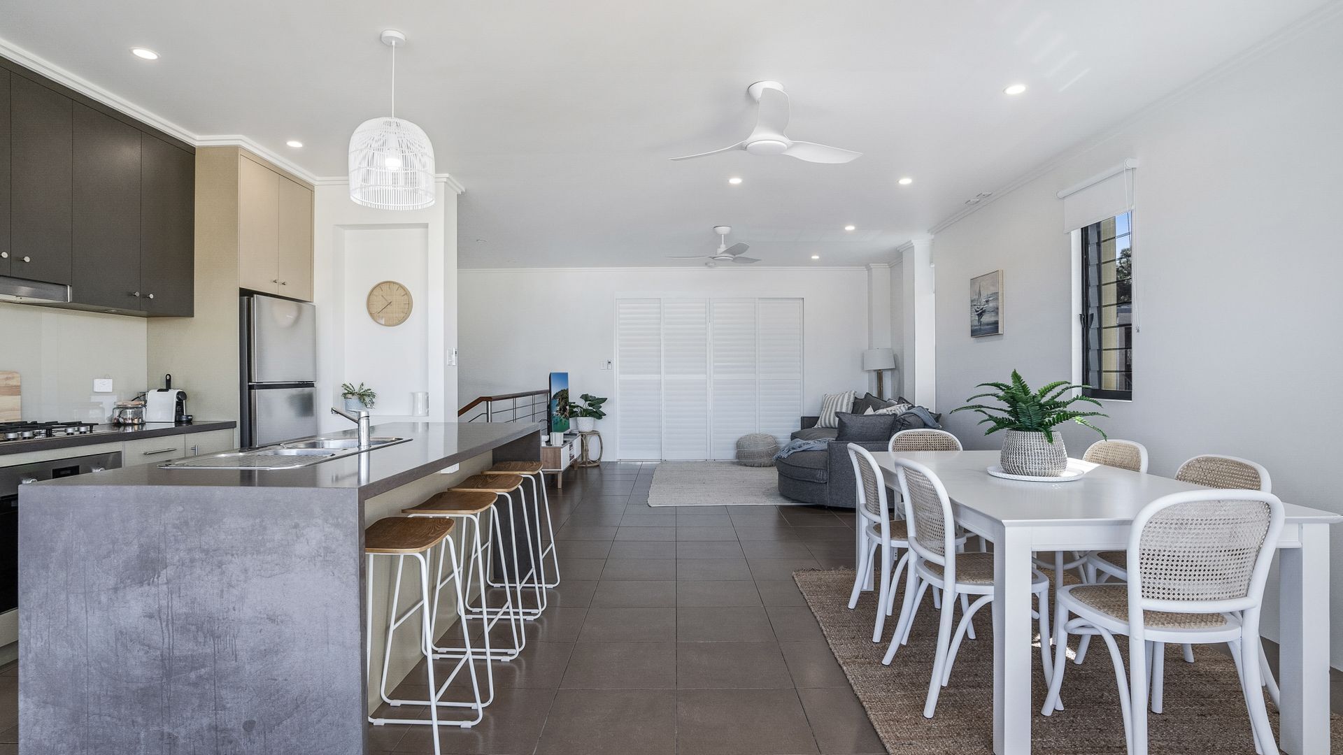 A modern kitchen with a central island, bar stools, a dining table with chairs, a ceiling fan, and a large window.
