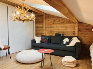 Um quarto aconchegante com um sofá cinza escuro, um banquinho branco arredondado, uma mesinha vermelha e teto de madeira.